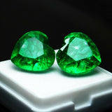 Natural green heart-shaped emerald pair positioned on top of a small white display box, with a dark black background adding contrast to highlight the gems’ rich color and depth.