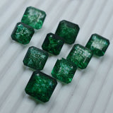 Nine emerald stones forming a triangle on grey fabric background – square cut, translucent green gems