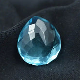 āFaceted blue pear-shaped gemstone placed on black fabric background.ā