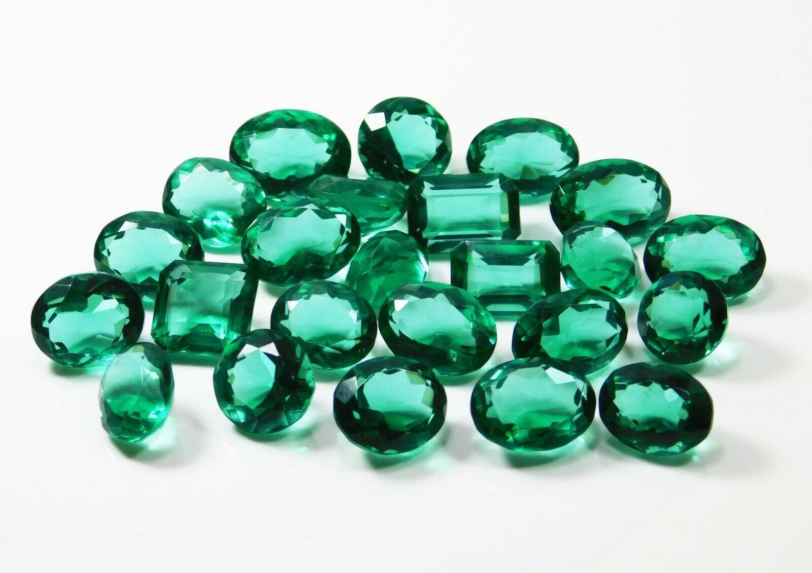 A set of lab-created green Muzo emeralds, including oval, circular, and rectangular shapes, arranged at the center in a white display, showcasing transparency and excellent cut.