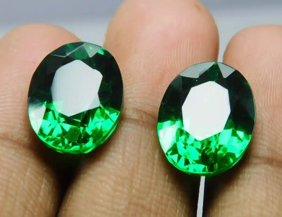 Two green gemstones held between fingers against a neutral background