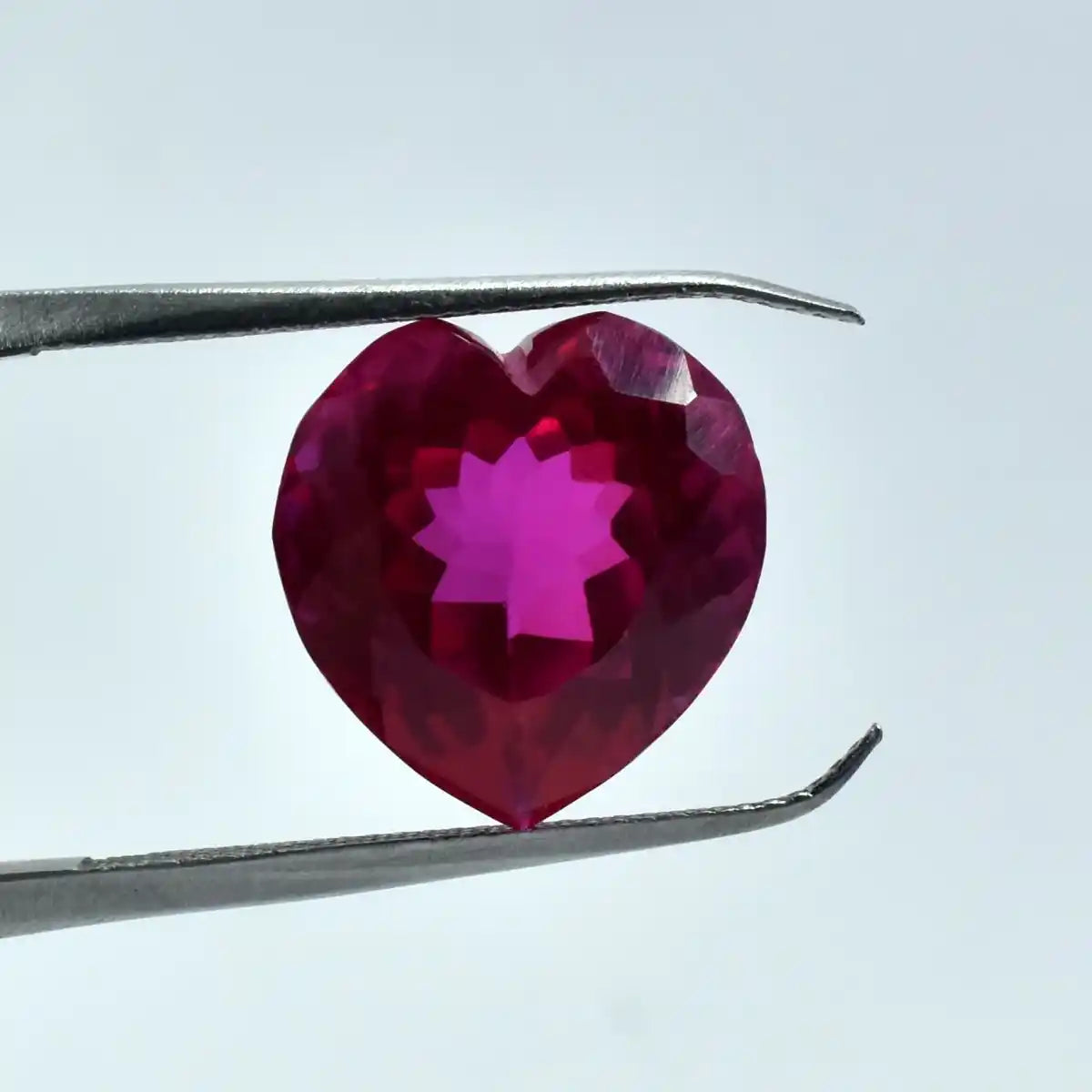 Heart-shaped red gemstone held by tweezers against a light blue background