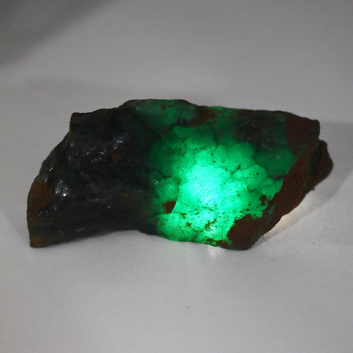 “Raw green gemstone with a bright internal glow, displayed on a white surface.”