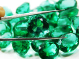 Close-up of a circular-shaped green lab-created emerald held by a jewel clamp with other emeralds softly blurred in the background, highlighting clarity and precision.